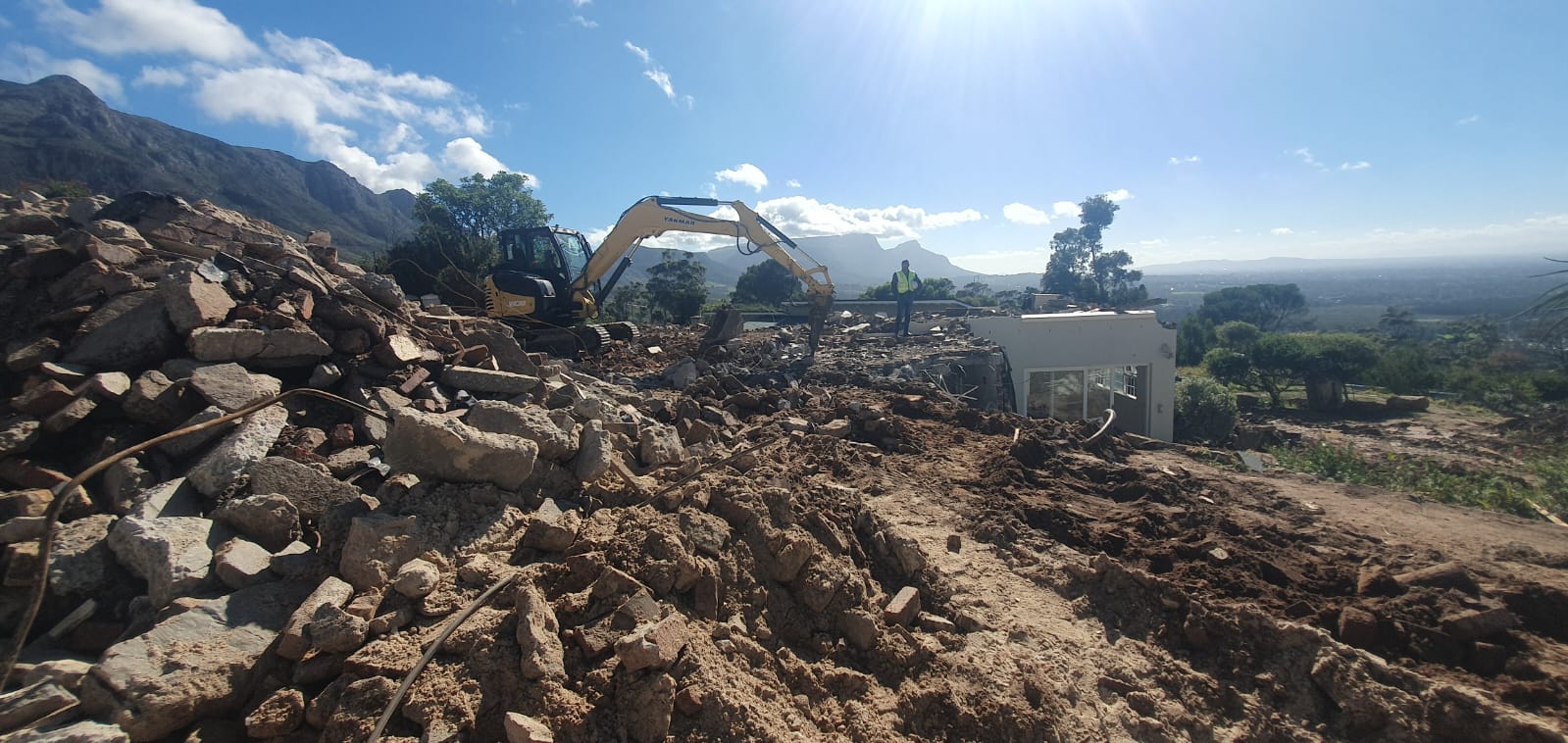 Excavator working through demolition rubble with mountain views behind